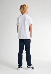 Hvid bomulds T-shirt, korte ærmer, rund hals, parret med mørkeblå jeans og hvide sneakers med sorte accenter, set fra ryggen.