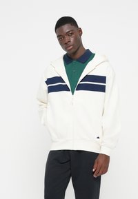 Biela mikina s kapucňou na zips, s námořníckymi a bielymi pruhmi cez hruď, má šnúrku na kapucni, pružné manžety a malé logo na boku.