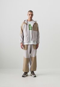 Gray and beige fleece jacket with large pockets, paired with matching trousers featuring a similar color scheme and a white shirt with a green logo.