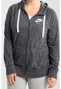 Šedá mikina na zip s bílými šňůrkami a logem Nike. Má dlouhé rukávy a dvě přední kapsy. Jemná textura s propleteným vzorem.