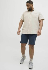 Beige Kurzarm-Hemd mit Knöpfen und strukturiertem Muster, kombiniert mit marineblauen Shorts. Das Modell trägt hellgraue Sneakers mit beigen Socken.