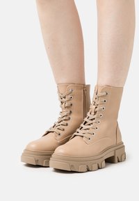 Bottes de combat en cuir beige avec une semelle épaisse, dotées d'œillets métalliques, de lacets épais et d'une fermeture éclair latérale pour un enfilage facile.