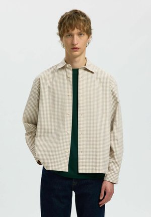 Ung mand iført beige ternet skjorte med knapper over grøn t-shirt og mørke jeans, står mod en ensfarvet lys baggrund.