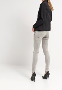 Žena stojící v světlých šedých skinny džínách, černých vysokých podpatcích a černém dlouhém rukávu, čelí doleva s rukama u kapes.