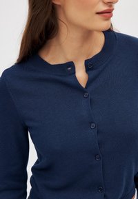 Frau, die eine dunkelblaue, knöpfbare Strickjacke mit rundem Ausschnitt trägt, bei der die obersten drei Knöpfe offen sind.