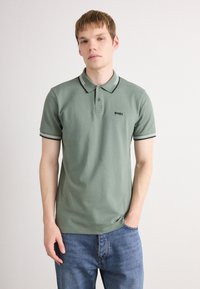 En ung mand står iført en salviegrøn polo-shirt med sort og hvid kant rundt om kraven og ærmerne, parret med blå jeans, imod en hvid baggrund.