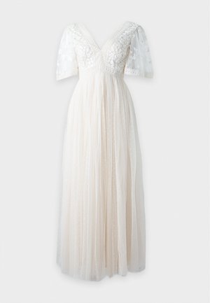 Femme portant une robe blanche longue jusqu’au sol avec un corsage en dentelle florale, des manches courtes transparentes et une jupe plissée à pois, chaussée de mules crème à talons.