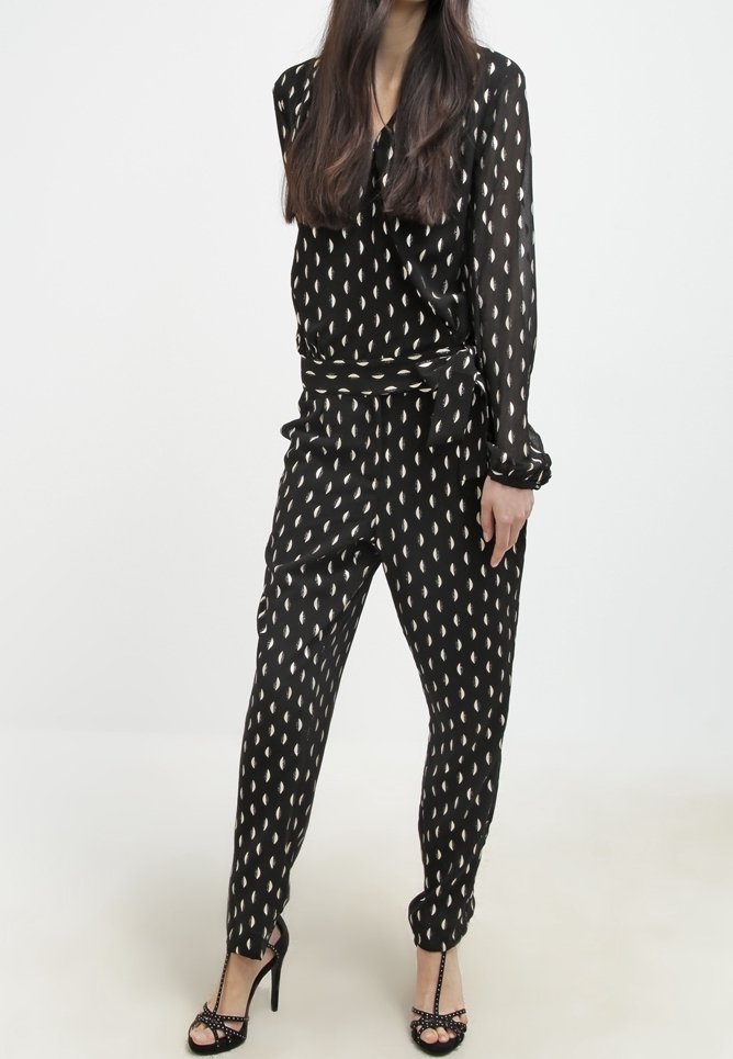 Femme portant une combinaison noire avec des motifs blancs en forme de feuilles, manches longues, taille nouée, et sandales noires à talons hauts à bout ouvert.