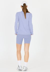 Kvinde med langt hår iført en hvid kasket, iført en lyse lilla langærmet top og matchende shorts, stående med ryggen mod kameraet.