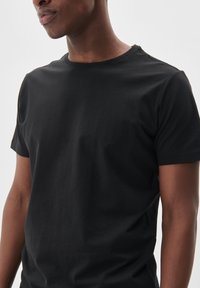 Tricou negru cu manșete scurte fabricat din bumbac, având un guler clasic și o textură fină. Designul este simplu și se mulează pe corp.