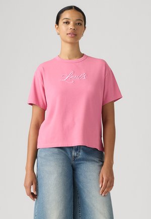 Γυναίκα που φοράει ροζ μπλουζάκι Levi's και ανοιχτό μπλε τζιν με φαρδιά μπατζάκια, στέκεται μπροστά σε ένα απλό ανοιχτό φόντο.
