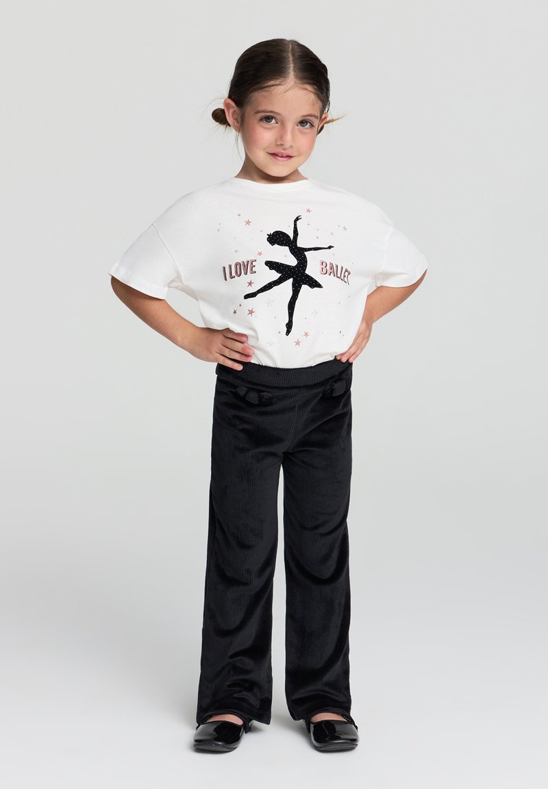 Biała koszulka z czarnym siluetą tancerki baletowej i błyszczącym tekstem, połączona z czarnymi spodniami w katedralne prążki i błyszczącymi czarnymi baletkami.