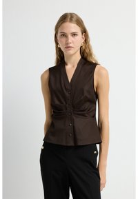 Braune ärmellose Bluse mit V-Ausschnitt, Knopfleiste und gefaltetem Taillendetail, kombiniert mit schwarzen Hosen mit goldenen Akzenten.