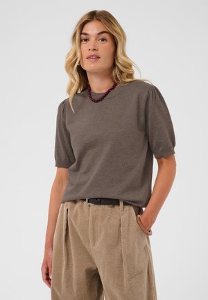 Femme portant un haut marron à manches courtes, un pantalon beige plissé en velours côtelé avec une ceinture, un collier de perles bordeaux et un bracelet marron.