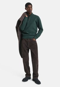 Groene pullover met half-rits kraag, gecombineerd met donkerbruine suède broek en zwarte schoenen. Een gestreepte jas over één arm gedrapeerd.