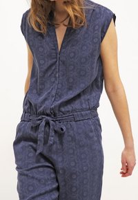 Mono sin mangas de color azul marino con sutiles patrones circulares, un escote en V y un lazo en la cintura, llevado por una persona de piel clara y cabello suelto.