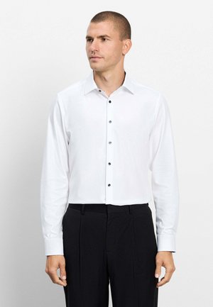 Mann trägt ein weißes Langarmhemd mit schwarzen Knöpfen und schwarze Hose, steht und sieht leicht zur Seite.