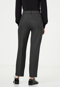 Osoba v uhlově šedých pruhovaných kalhotách a černých lesklých oxfordkách, stojící před jednoduchým bílým pozadím.