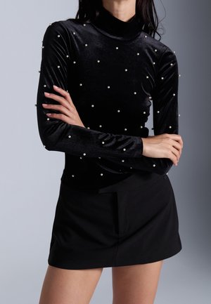 Femme portant un haut en velours noir à manches longues avec des perles blanches et une mini-jupe noire, bras croisés devant.