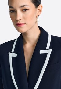 Blazer azul marino con solapas blancas y detalle en el bolsillo en contraste, con un ajuste entallado y hombros estructurados. Textura de tela suave.