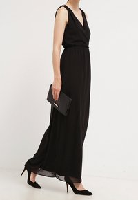 Czarna, bezrękawna sukienka maxi z głębokim dekoltem w kształcie litery V, marszczoną talią i zwiewnym dołem. Noszona z czarnymi szpilkami i czarną kopertówką.