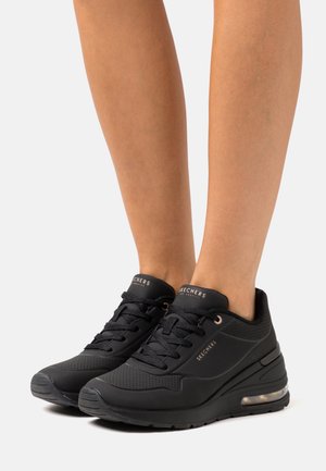Benen met zwarte Skechers vetersneakers met gedempte zolen, zichtbare luchtkamer en merklogo aan de zijkant en op de tong.