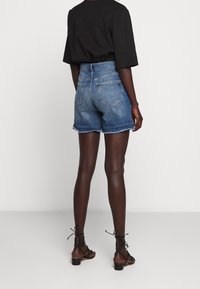 Person som bär blå högmidjade jeansshorts med fransiga kanter, en lös svart t-shirt och svarta snörskor, står mot en vit bakgrund.