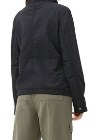 Veste noire légère avec un col montant, coutures visibles, taille élastique et poignets ; associée à un pantalon cargo vert olive.