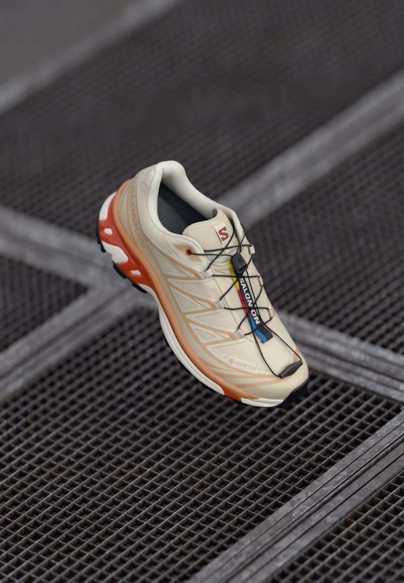 Bež tenisica za trčanje s narančastim detaljima, dizajnirana s teksturiranim gornjim dijelom, crnim potplatom i integriranim vezicama. Sadrži GORE-TEX oznaku.