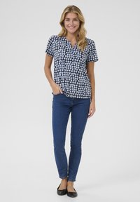 Korte mouw marineblauwe blouse met een lichtblauw vierkant patroon, gecombineerd met strakke blauwe jeans en zwarte ballerina's. Model poseert met één hand in de zak.