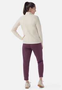 Giacca zip-up beige chiaro con colletto alto e silhouette aderente, abbinata a pantaloni attillati viola scuro e sneakers neutre.
