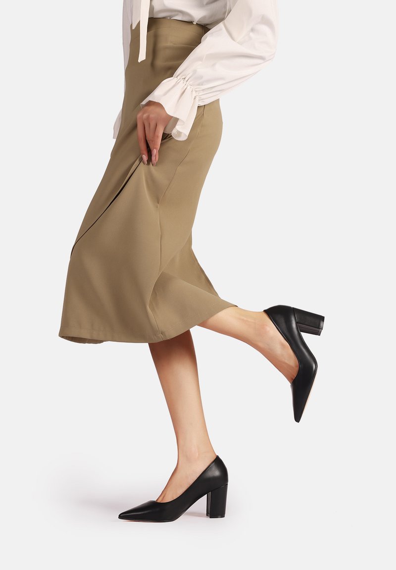 Scarpe nere in pelle con tacco alto, punta affusolata e tacco a blocco indossate su un piede. Abbinato a una gonna chiara e una camicia bianca.