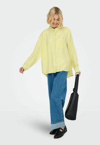 Helle gelbe Langarmbluse mit einer Tasche, kombiniert mit lockeren blauen Jeans. Das Modell hält eine schwarze Tasche und trägt schwarze Slipper.