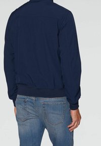 Mørkeblå bomberjakke med ribbet elastisk talje og ærmer, glat tekstur og et enkelt design, parret med falmede blå jeans.