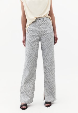 Kvinde iført grå vidbenede bukser med høj talje og diskret zebra print, kombineret med en cremefarvet top og sorte åbne hæle.