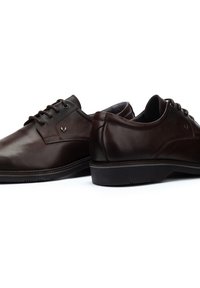 Chaussures en cuir marron avec un bout arrondi, un laçage, un logo discret, et des accents texturés contrastants sur le talon.