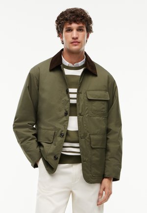 Mand med krøllet hår iført en olivengrøn jakke med sorte knapper over en grøn og hvidstribet sweater og hvide bukser.