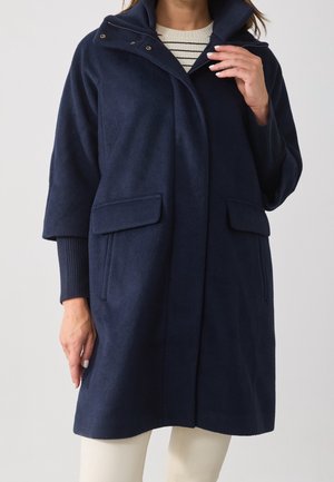 Kobieta ubrana w granatowy płaszcz z kieszeniami z klapkami i szerokimi rękawami o długości do łokcia, nałożony na biało-czarną pasiastą bluzkę.