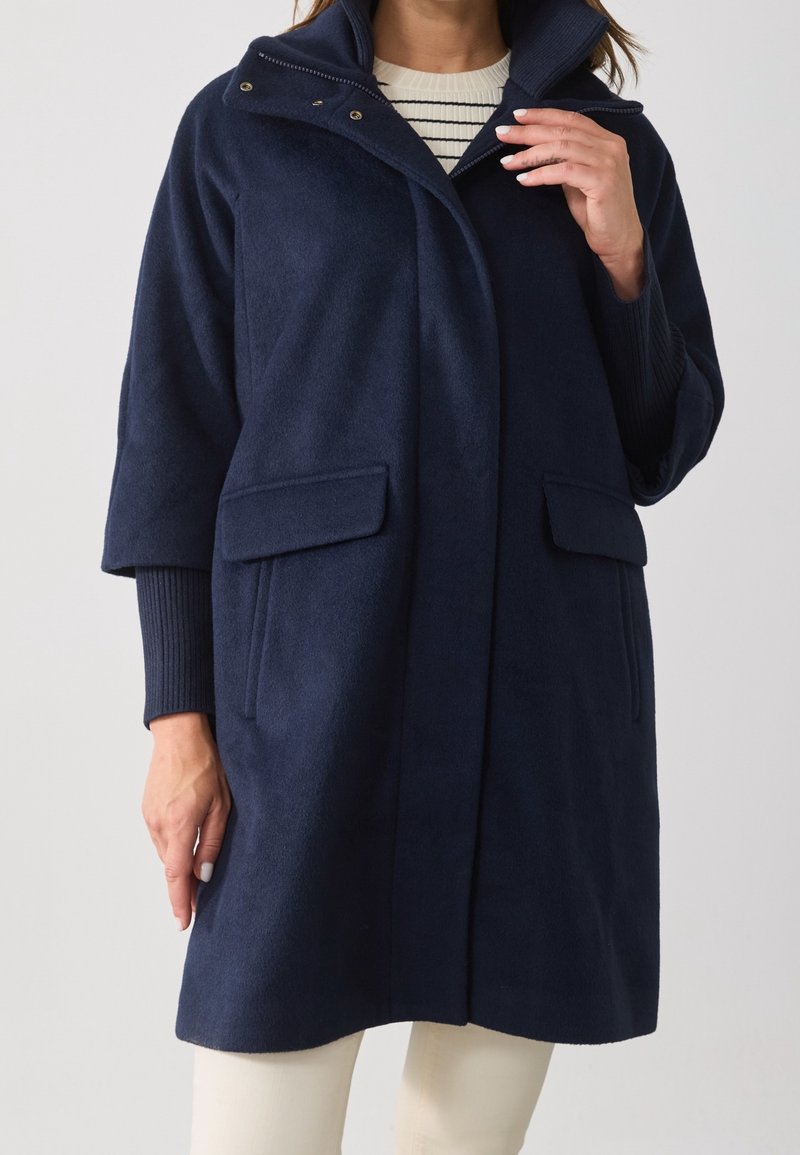 Femme portant un manteau bleu marine avec poches à rabat et larges manches trois-quarts par-dessus un haut rayé blanc et noir.
