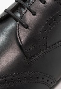 Chaussure habillée en cuir noir avec un motif brogue détaillé, un bout rond et des lacets plats. Présente une texture lisse et des coutures contrastées.