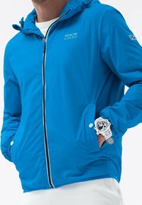 Blå letvægtsjakke med lynlåslukning foran, der har hætte, elastiske manchetter og et lille hvidt tekstmotiv på brystet.