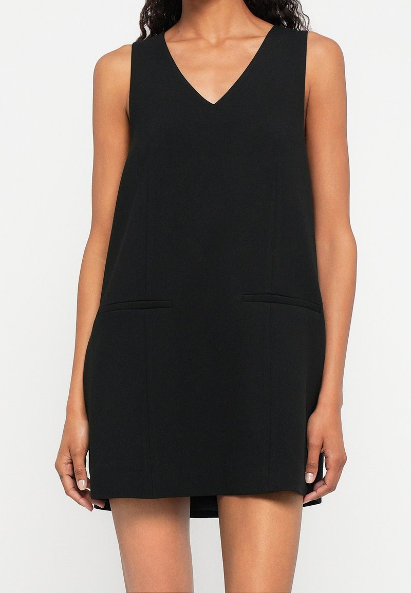 Femme portant une mini-robe noire sans manches à col en V avec deux poches fendues horizontales à l'avant.