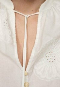 Bílá blůza s květinovou výšivkou na hrudi, s hlubokým výstřihem do V, zavazováním a dřevěnými knoflíky. Jemný, lehký materiál.