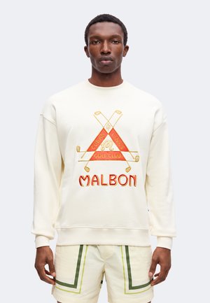Mann trägt cremefarbenen Sweatshirt mit rotem und goldenem "Malbon Cuban Links Golf Club" Logo und cremefarbene Shorts mit grünen Streifen, steht vor einfachem Hintergrund.