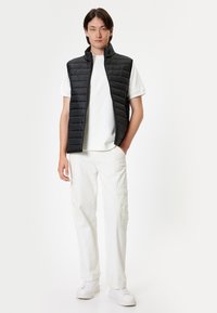 Gilet imbottito nero con colletto alto, design trapuntato testurizzato, indossato sopra una maglietta bianca e abbinato a pantaloni cargo bianchi e sneaker bianche.