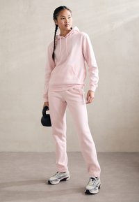 Sweatshirt rosa claro com bolso frontal, calças de treino rosa a condizer e um kettlebell preto. Os ténis têm detalhes em cinza e roxo.