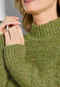 Grüner Strickpullover mit hohem Rippenkragen. Die Textur ist grob und besteht aus einem Mix aus hellen und dunklen grünen Fasern, die Tiefe erzeugen.
