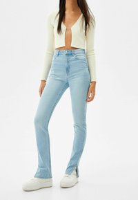 Maupert blue jeans dengan pinggang tinggi dan garis samping di bagian pergelangan kaki, terbuat dari denim, dipadukan dengan atasan putih lengan panjang berkain ribbed yang dipotong pendek.