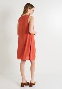 Vestido sin mangas de color naranja con una tela texturizada, de longitud hasta la rodilla, corte holgado, detalle de pliegue en la parte trasera, combinado con zuecos de plataforma marrones.