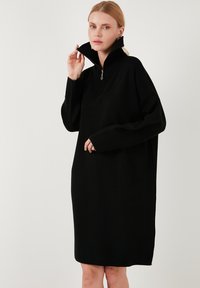 Rochie neagră supradimensionată cu guler înalt și fermoar la jumătate. Dispune de mâneci lungi și o croială lejeră, fiind fabricată dintr-un material moale și texturat.